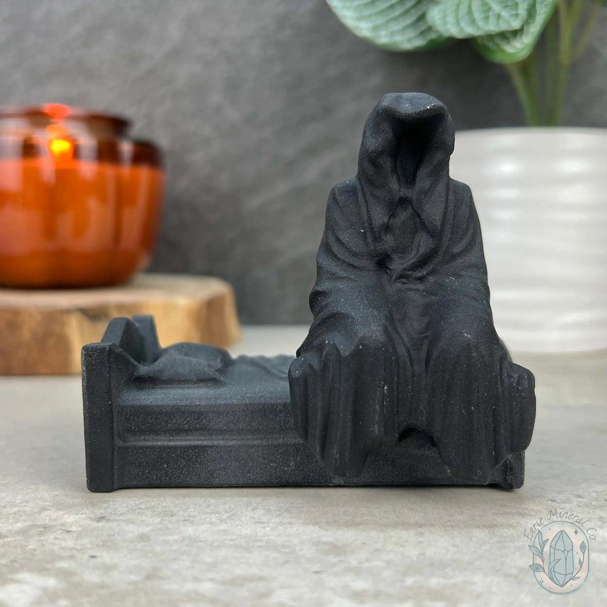 An obsidian grim reaper figurine of a hooded seated figure on a small bed, displayed on a stone surface with a blurred amber candle and green plant in the background.