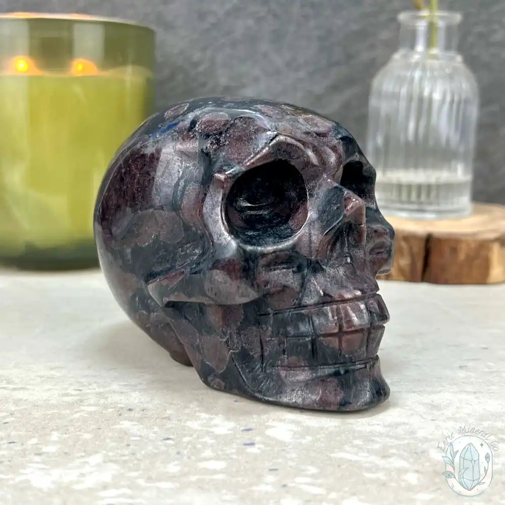 A polished Garnet crystal skull carving with black, gray, and deep purple marbling, displayed on a light stone surface with a green candle and a glass bottle in the background.
